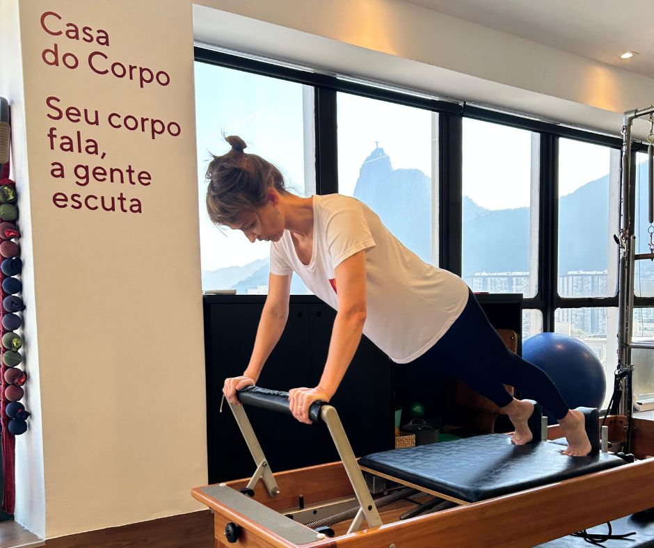 Aluna em exercício de pilates no reformer, com o Corcovado visível pela janela — Casa do Corpo, Botafogo